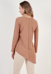 Maglia marrone a maniche lunghe, a coste, con orlo asimmetrico, caratterizzata da una cucitura nella parte posteriore. Indossata con pantaloni beige, dal tessuto liscio.