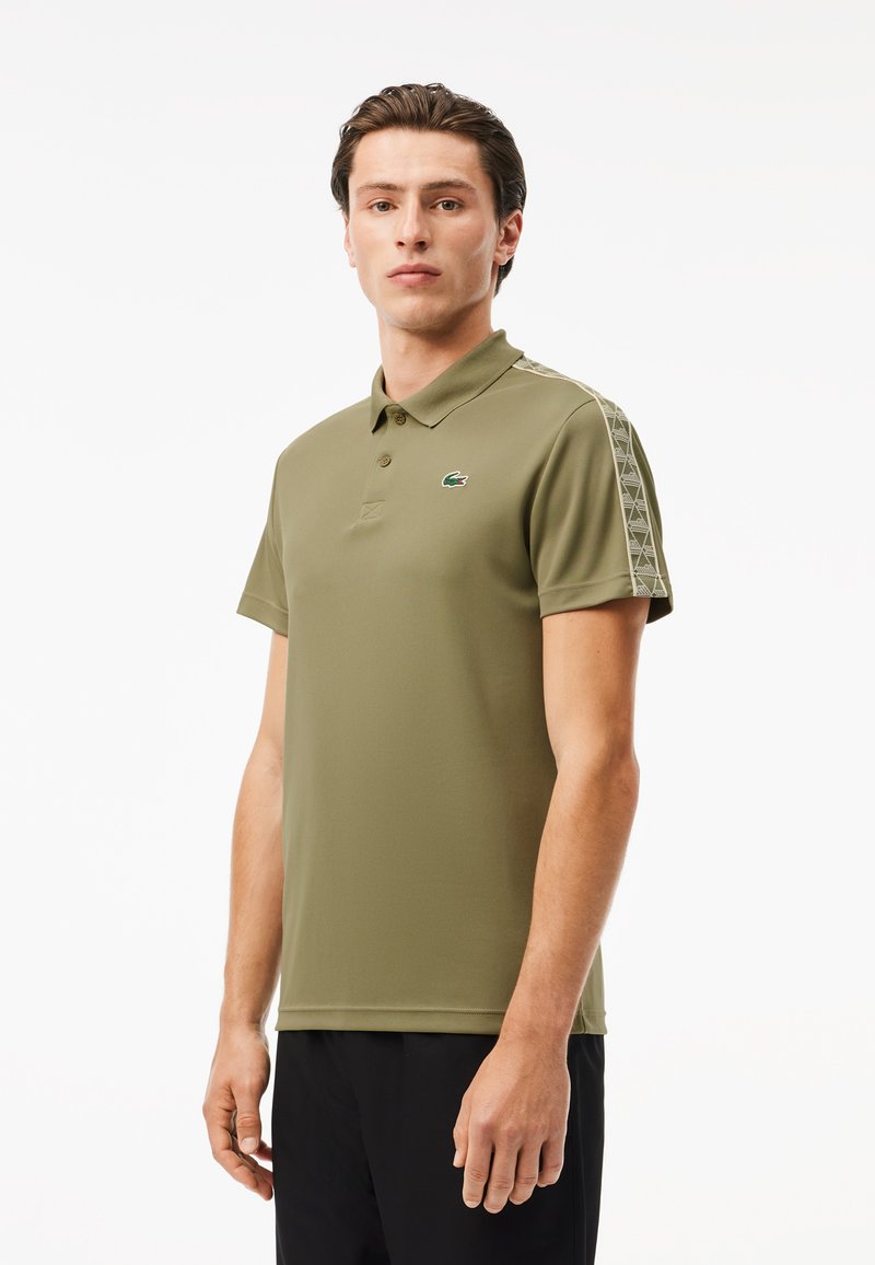 Polo vert olive à manches courtes et col boutonné. Présente des motifs géométriques le long des épaules et un petit logo sur la poitrine.