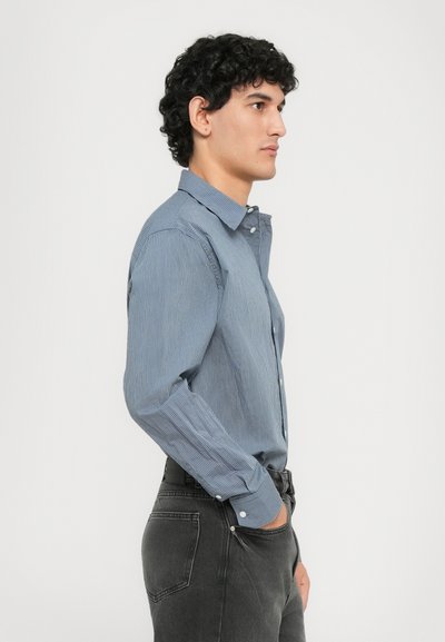 Jeune homme aux cheveux noirs bouclés portant une chemise à rayures bleues et un jean noir, se tenant de profil avec une main dans la poche.