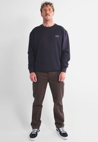 Sweat-shirt marine avec un petit logo sur la poitrine, associé à un pantalon marron et des baskets noires avec des semelles blanches et un motif texturé.