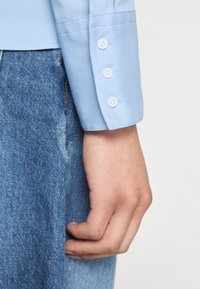 Main droite reposant à côté d'un jean en denim bleu, portant une manche de chemise bleu clair avec trois boutons blancs sur le poignet.