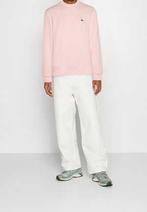 Man wearing light pink long-sleeve sweatshirt, white wide-leg pants, and gray-green athletic shoes, standing against a plain background.