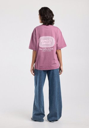 Persoon met kort donker haar in een loszittend roze T-shirt met de tekst "Sunset Drive" en een brede blauwe spijkerbroek, staand met de rug naar de kijker.