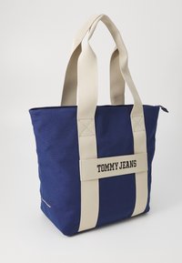 Mörkblå canvas shoppingväska med krämfärgade remmar och en logotypapplikation som säger "TOMMY JEANS", med dragkedja och horisontell form.