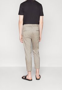 Persoon in beige cropped broek, zwart shirt met korte mouwen en zwarte slipper-sandalen, staande tegen een effen lichte achtergrond.