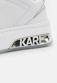 Białe skórzane sneakersy z metalicznym logo na pięcie, z teksturowanym chromowanym detalem i kontrastującą szaro-skalową podeszwą.