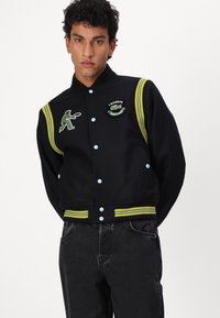 Veste varsity noire avec des boutons bleus, des poignets rayés vert et jaune et des applications brodées de joueur de tennis et de logos.