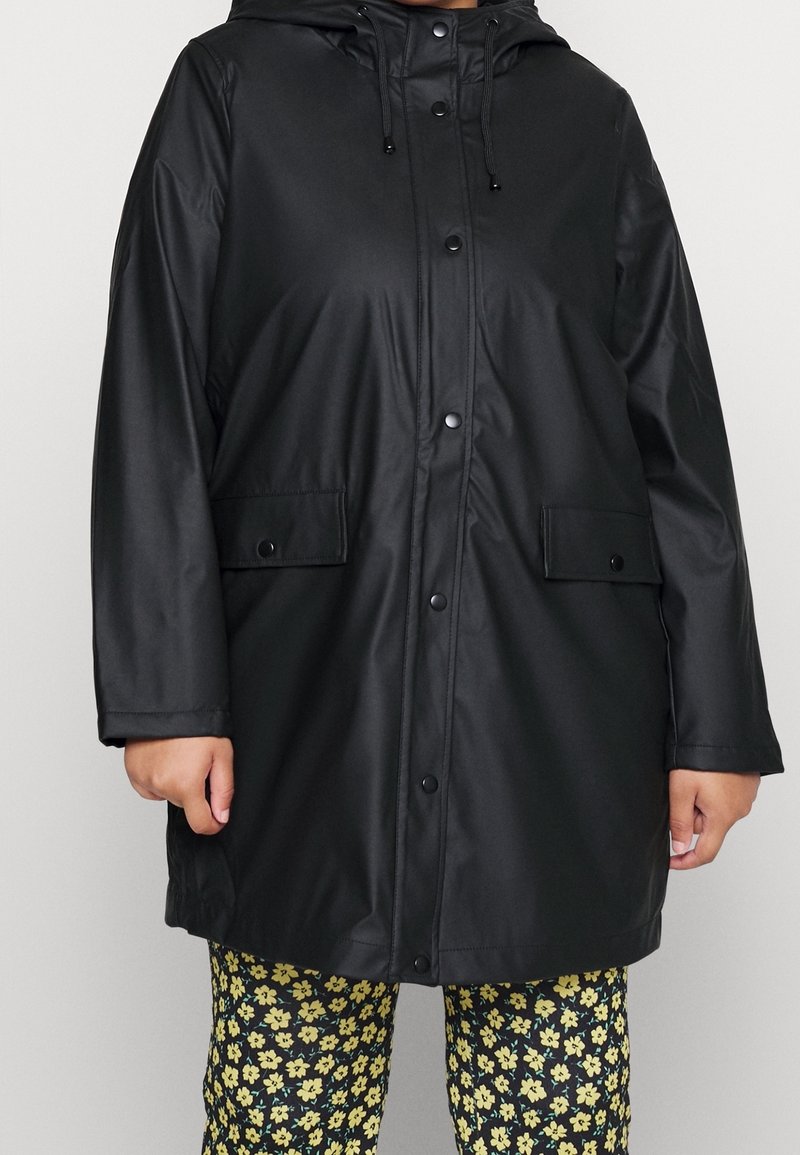 Personne portant un imperméable noir à capuche avec des boutons-pression à l'avant et des poches à rabat, associé à un pantalon noir à motif floral jaune.