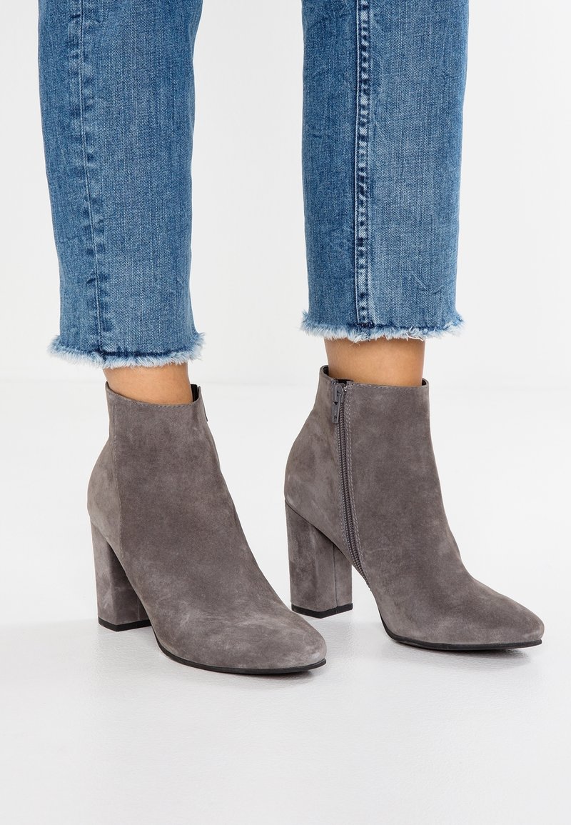 Bottines en daim gris avec un talon bloc haut et une fermeture éclair latérale, associées à un jean bleu à ourlet effiloché. Texture lisse et design à bout pointu.