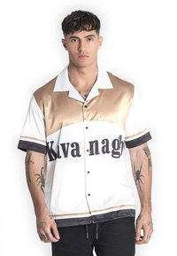 Camisa de manga corta en blanco, dorado y negro; acabado de satén suave con letras en negrita "Kva nagh" y acentos a rayas en las mangas y el dobladillo.