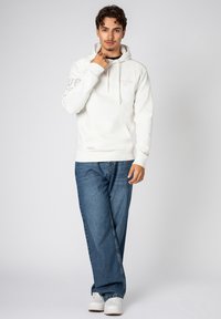 White hoodie with a kangaroo pocket, adjustable drawstring hood, and logo details on sleeves. Paired with blue wide-leg jeans and white sneakers.