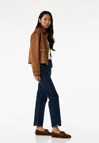 Chaqueta de ante marrón corta, blusa crema, vaqueros rectos azul oscuro y zapatos de ante marrón con detalles de cordones. Modelo se encuentra de lado.
