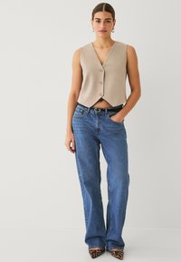 Beige kurz geschnittene Weste mit V-Ausschnitt und Knopfverschluss, kombiniert mit blauen Schlagjeans. Modell trägt Leopardenmusterschuhe und hat die Hände in den Taschen.