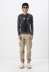 Gray long-sleeve shirt with logo, beige cargo pants with ankle cuffs, black crossbody bag, and round sunglasses. White sneakers with brown soles.