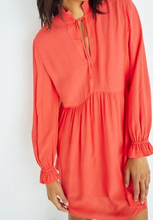 Femme portant une robe corail à manches longues avec une taille froncée, des poignets à volants et un lien à l’encolure sur fond blanc.
