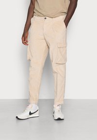 Beiga kordelbyxor i cargo-modell med avslappnad passform, med sidofickor, knappstängning och sydda detaljer. Bärs med vita sneakers.