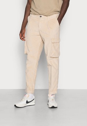 Beige corduroy cargobroek met een relaxed fit, voorzien van zijzakken, een knoopsluiting en gestikte details. Gedragen met witte sneakers.