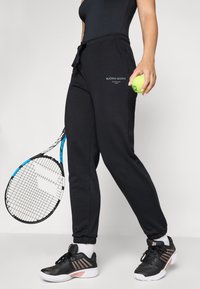 Svarta sweatshirtbyxor med dragsko i midjan, med en tryckt logotyp. I kombination med svarta sportskor och håller en tennisboll.