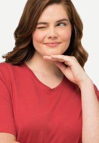 Červené tričko s výstřihem do V vyrobené z jemného materiálu, s krátkými rukávy a volným střihem. Modelka má vlnité vlasy a opírá ruku o bradu.