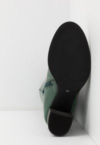 Botte en cuir vert avec une semelle en caoutchouc noire texturée, affichée avec le talon vers le haut. Taille 37 indiquée sur la semelle.