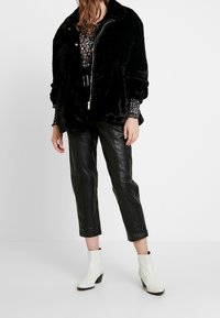 Svart fuskpälsjacka med krage, dragkedjestängning och rynkade ärmar; kombinerad med högmidjade svarta läderbyxor och vita ankelstövlar.