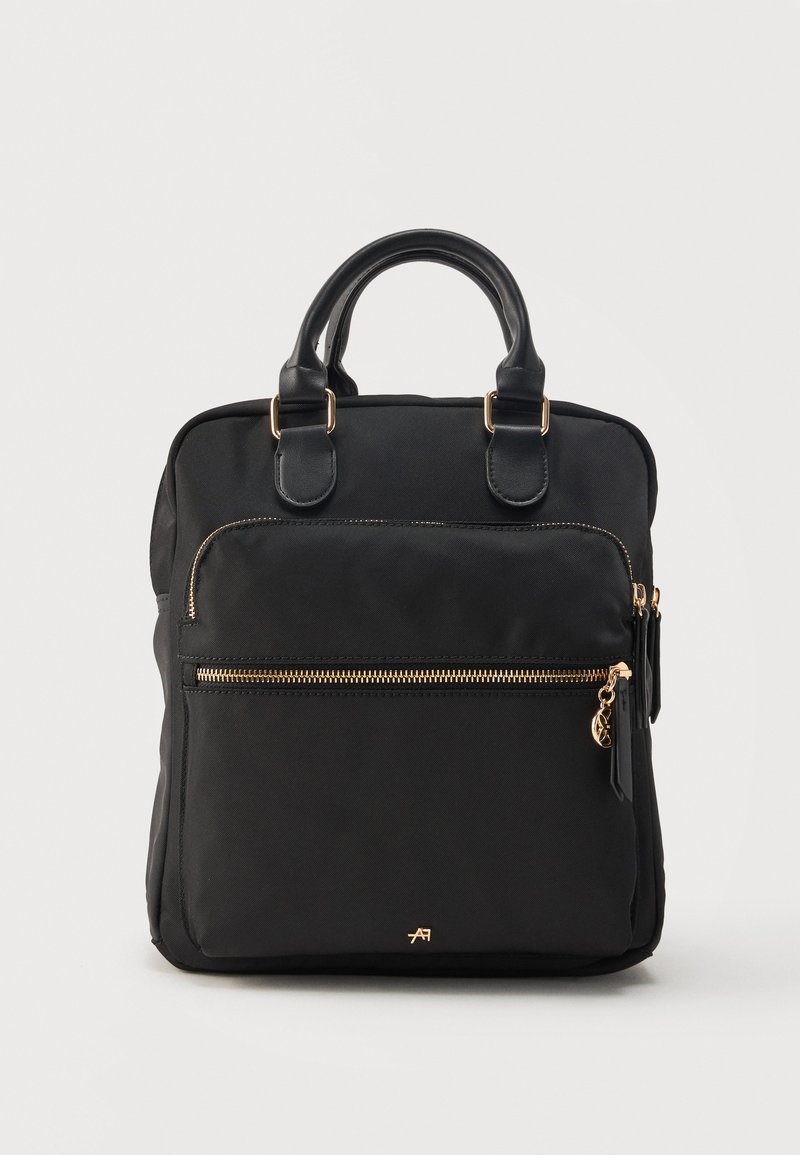 Sac à dos rectangulaire noir avec deux anses, poche avant zippée avec fermeture éclair dorée et tirette, et petit logo de marque doré en bas au centre.