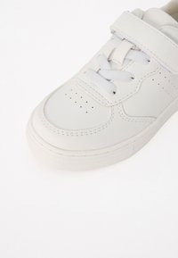 Baskets blanches avec lacets et une sangle auto-agrippante, comportant des détails perforés au niveau des orteils et sur le côté, sur un fond blanc uni.