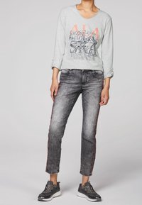 Světlé šedé tričko s dlouhým rukávem a grafickým potiskem, spárované se šedými roztrhanými džíny s červenými bočními pruhy a černými sportovními teniskami.