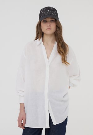 Camicia bianca, oversize, a maniche lunghe in tessuto leggero con chiusura a bottoni e colletto. Abbinata a un berretto scuro con borchie.