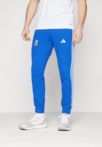 Blue athletic pants with white Adidas stripes and Real Madrid logo. Made of soft fabric, featuring a tapered design and elastic cuffs.