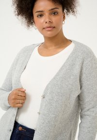 Lichtgrijze cardigan van zacht breimateriaal met een V-hals en vijf knoopsluitingen, gedragen over een witte, aansluitende t-shirt.