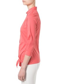 Camisa de manga larga color coral con cuello, detalle de corbata en la cintura, tejido suave y silueta ajustada. Llevada con pantalones blancos.