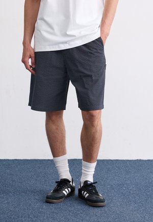 Person iført sorte teksturerede shorts, hvide crew-sokker, sorte og hvide sneakers og en hvid T-shirt, der står på blåt tæppe.