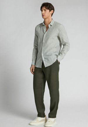 Mężczyzna ubrany w zielono-białą koszulę w pionowe paski na guziki, ciemnozielone spodnie i białe trampki, stoi z jedną ręką w kieszeni.