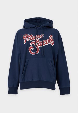 Femme debout en hoodie Marc Jacobs bleu marine, mini-jupe en jean bleu à motifs et ballerines argentées sur fond blanc uni.