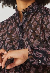 Blusa negra transparente con patrones de helechos en rosa, puntos texturizados y un cuello con volantes. Cerrada con botones y puños fruncidos.