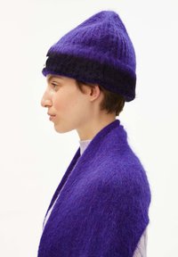 Person mit lila Strickmütze und passendem flauschigen Schal, die nach links auf einem schlichten weißen Hintergrund blickt.