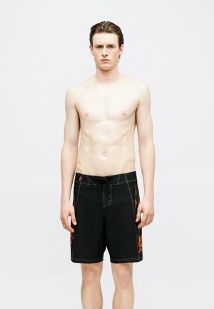 Junger Mann mit hellem Teint und nassen Haaren steht oberkörperfrei in schwarzen Boardshorts mit orangefarbenen Flammenmustern an den Seiten vor einem schlichten weißen Hintergrund.