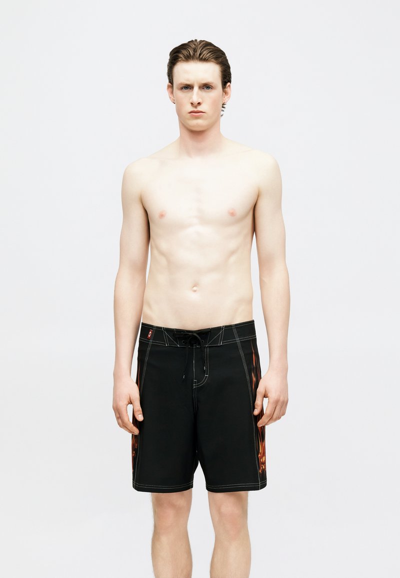 Νέος άνδρας με ανοιχτόχρωμη επιδερμίδα και βρεγμένα μαλλιά, στέκεται γυμνόστηθος φορώντας μαύρο σορτς για σερφ με πορτοκαλί φλόγες στα πλαϊνά, μπροστά σε απλό λευκό φόντο.