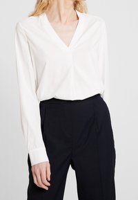 Blouse blanche à manches longues, encolure en V, avec un léger éclat. Associée à un pantalon sombre et ajusté. Tissu lisse et coupe décontractée.