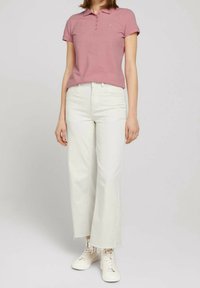 Rosa Poloshirt mit kurzen Ärmeln, weiße Jeans mit weitem Bein und cremefarbene Sneakers. Schlichte Knöpfe am Shirt und ausgefranster Saum an der Jeans.