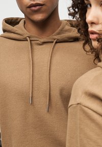 Καφέ υφασμένη φούτερ με κουκούλα, κορδόνια με μεταλλικές άκρες, σε άνετη εφαρμογή και ορατές ραφές στους ώμους.