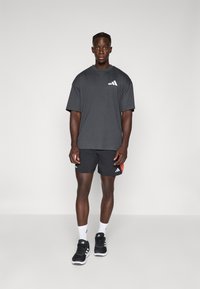 Homme se tenant contre un fond uni, portant un t-shirt Adidas gris foncé, un short noir avec des détails orange, des chaussettes blanches et des baskets Adidas noires.