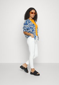 Camisa oversized com padrão floral azul sobre uma blusa laranja brilhante, combinada com jeans brancos justos e sandálias de plataforma pretas. Óculos de sol laranja.