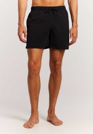 Homme portant un short de bain noir avec taille élastique et cordon, debout pieds nus devant un fond clair uni.