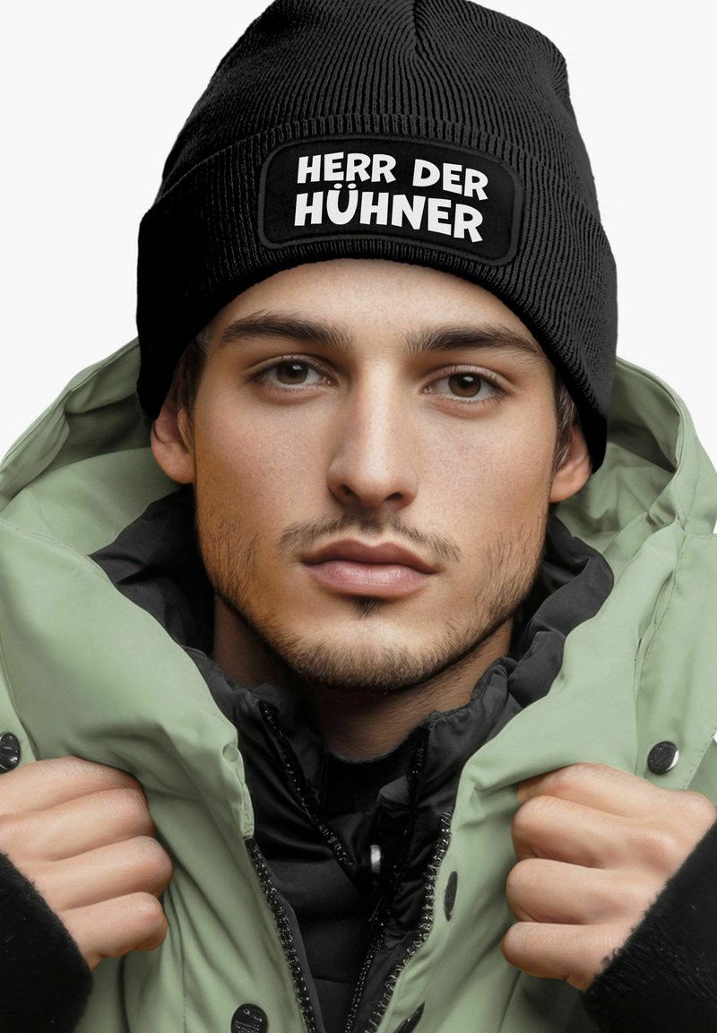 Black ribbed beanie with "HERR DER HÜHNER" text, paired with a quilted olive green jacket, featuring a high collar and black accents.