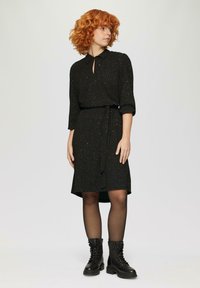 Schwarzes Kleid mit kragenförmigem Ausschnitt, Schlüsselöffnungsdetails, tailliertem Design und drei Viertel langen Ärmeln. Hat einen dezenten Glitzereffekt und endet knielang.