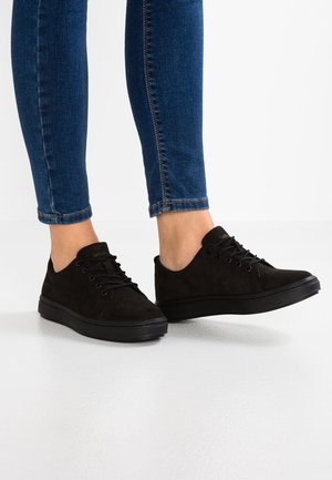 Jambes en jean skinny bleu portant des baskets basses à lacets noires, debout sur un sol blanc devant un fond blanc.