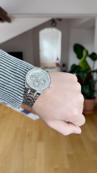 Montre en métal avec un boîtier octagonal, texture argentée, trois sous-cadrans et un bracelet maillon. Portée sur une manche rayée contre un sol en bois.
