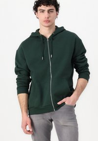 Hoodie zippé vert foncé avec une poche kangourou, des poignets ajustés et une capuche avec cordon de serrage, porté avec un jean gris clair.